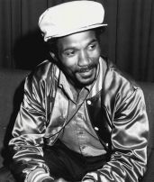 Portrait of Greg Richardson, seated, wearing an unbuttoned satin jacket and wool cap.
